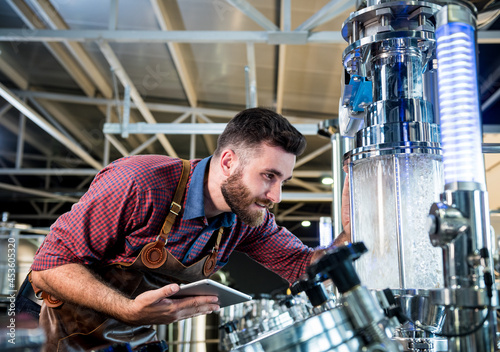 Young male brewer in leather apron supervising the process of beer fermentation at modern brewery factory