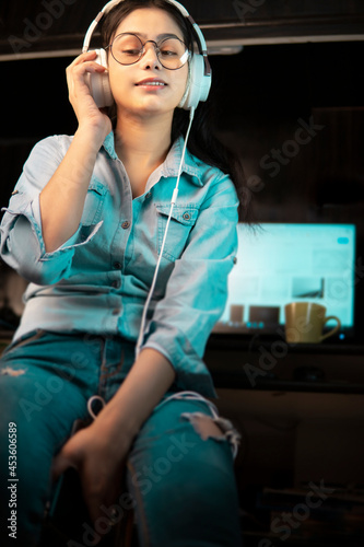 Low angle image of Beautiful happy bespectacled Asian, Indian young woman in denim shirt sitting at desktop and listening music through headphones while taking break from work at home. 