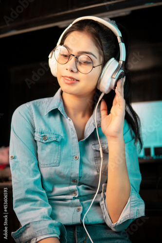 Low angle image of Beautiful happy bespectacled Asian, Indian young woman in denim shirt sitting at desktop and listening music through headphones while taking break from work at home. 
