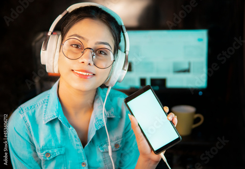 Beautiful happy bespectacled Asian, Indian young woman sitting at desktop at home and listening music through headphones and showing mobile screen to the camera with the toothy smile at home.