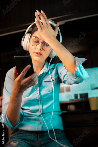 Low angle image of Beautiful bespectacled Asian, Indian young woman in denim shirt sitting at a desktop and receiving bad news on the phone while listening music through headphones at home.