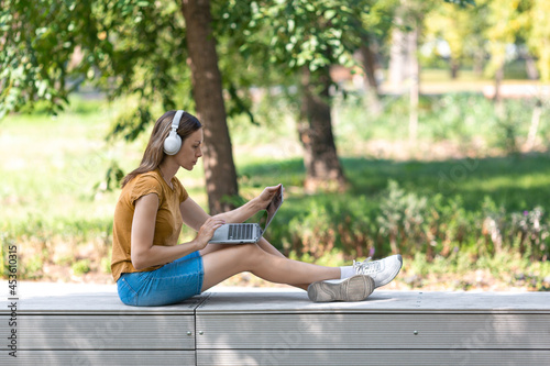 Digital nomad. Caucasian woman in wireless headphones sits on a parapet and working on a laptop in the public park outdoor in summer, side view, selected focus. © junky_jess