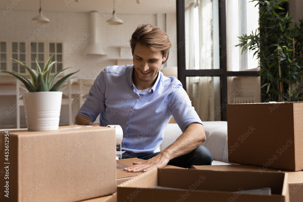 Smiling young handsome man using adhesive tape packing belongings in big carton boxes, feeling excited of moving into new apartment. Happy millennial homeowner preparing things for removal service. Smiling young handsome man using adhesive tape packing belongings in big carton boxes, feeling excited of moving into new apartment. Happy millennial homeowner preparing things for removal service.