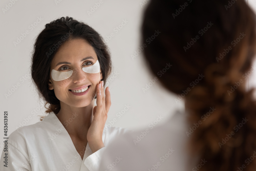 Head shot attractive millennial hispanic woman in bathrobe with applied anti wrinkles patches looking at mirror, moisturizing under eyes area, improving elasticity, enjoying facial skincare spa. Head shot attractive millennial hispanic woman in bathrobe with applied anti wrinkles patches looking at mirror, moisturizing under eyes area, improving elasticity, enjoying facial skincare spa.