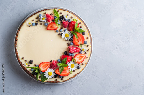 Cheesecake pie with fresh berries, strawberries, blueberries and blackberries and mint chamomiles in a round plate on a grey background.