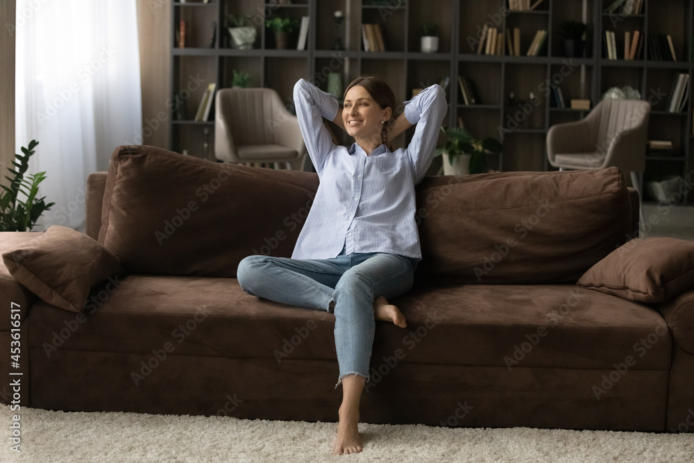 Full length smiling woman relaxing on couch at home, happy dreamy beautiful young female leaning back with hands behind head, visualizing good future, new opportunities, enjoying weekend Full length smiling woman relaxing on couch at home, happy dreamy beautiful young female leaning back with hands behind head, visualizing good future, new opportunities, enjoying weekend