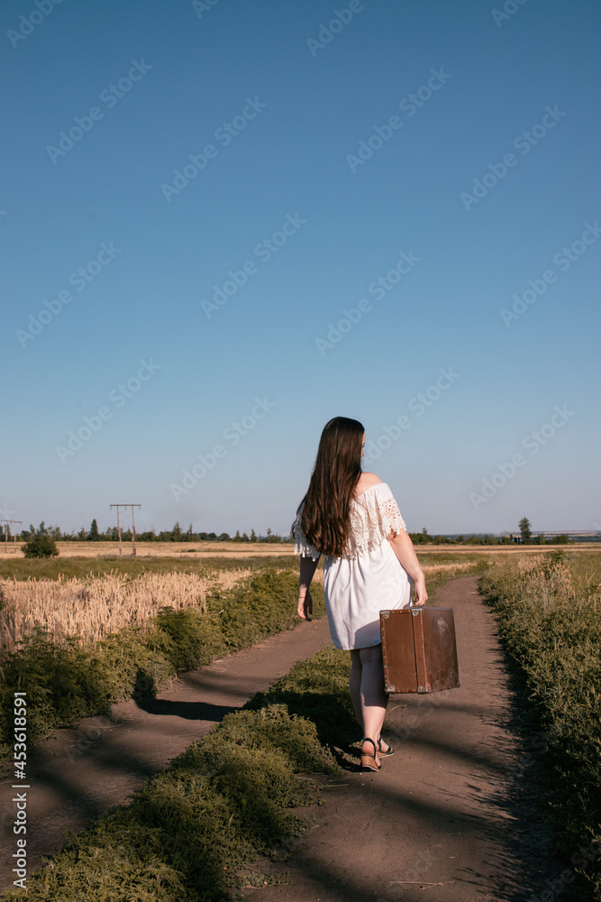 Beautiful girl in a white dress with a suitcase in a field of golden wheat. Journey to the 80s.