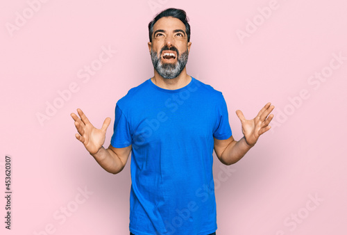 Middle aged man with beard wearing casual blue t shirt crazy and mad shouting and yelling with aggressive expression and arms raised. frustration concept.