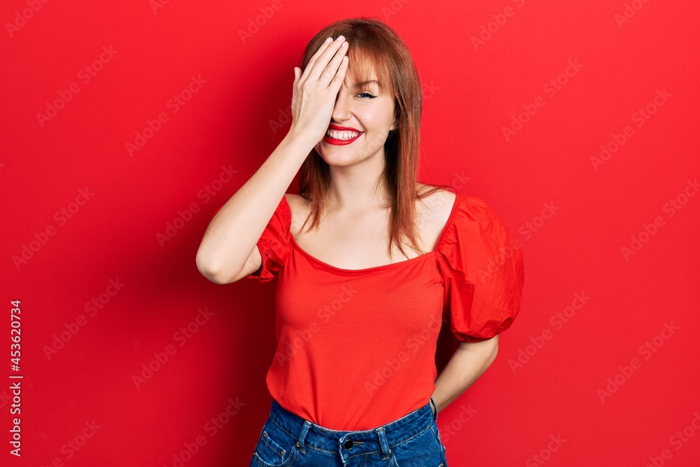 Redhead young woman wearing casual red t shirt covering one eye with hand, confident smile on face and surprise emotion. Redhead young woman wearing casual red t shirt covering one eye with hand, confident smile on face and surprise emotion.