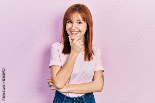 Redhead young woman wearing casual pink t shirt looking confident at the camera smiling with crossed arms and hand raised on chin. thinking positive.