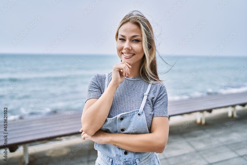 Young caucasian woman outdoors looking confident at the camera smiling with crossed arms and hand raised on chin. thinking positive.