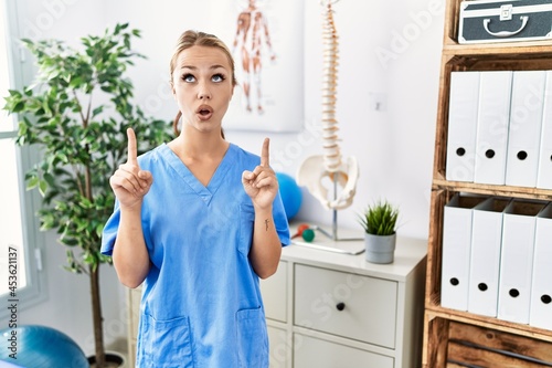Young caucasian woman working at pain recovery clinic amazed and surprised looking up and pointing with fingers and raised arms.