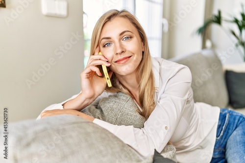 Young blonde woman talking on the smartphone sitting on the sofa at home.