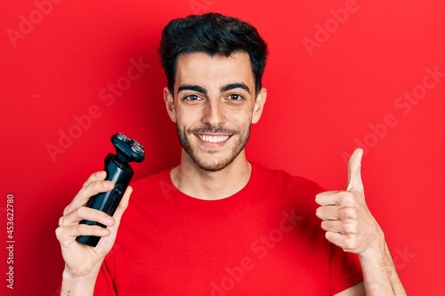 Young hispanic man holding electric razor machine smiling happy and positive, thumb up doing excellent and approval sign