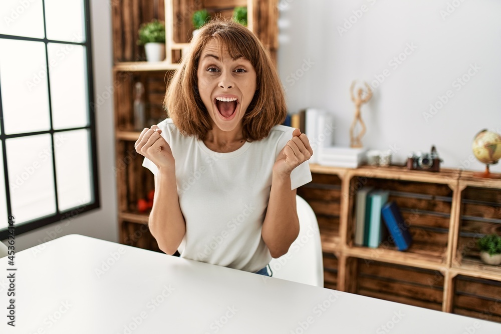 Young caucasian girl wearing casual clothes sitting on the table at home celebrating surprised and amazed for success with arms raised and open eyes. winner concept. Young caucasian girl wearing casual clothes sitting on the table at home celebrating surprised and amazed for success with arms raised and open eyes. winner concept.