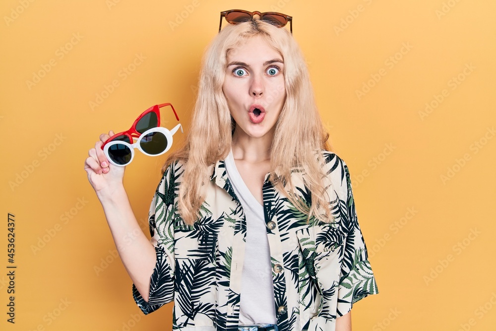 Beautiful caucasian woman with blond hair holding many sunglasses scared and amazed with open mouth for surprise, disbelief face Beautiful caucasian woman with blond hair holding many sunglasses scared and amazed with open mouth for surprise, disbelief face