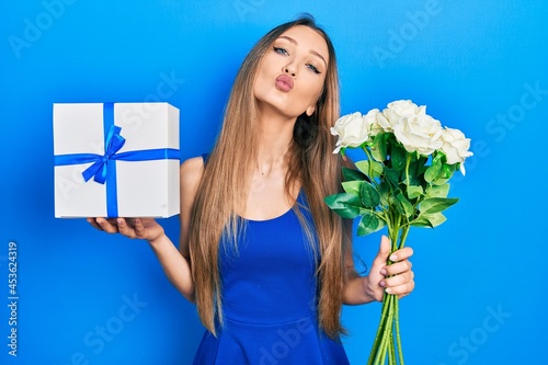 Young blonde girl holding anniversary present and bouquet of flowers looking at the camera blowing a kiss being lovely and sexy. love expression.
