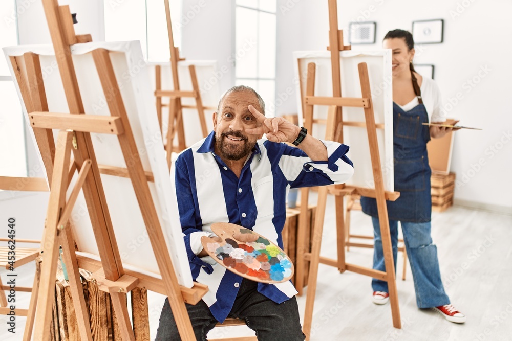 Senior artist man at art studio doing peace symbol with fingers over face, smiling cheerful showing victory Senior artist man at art studio doing peace symbol with fingers over face, smiling cheerful showing victory