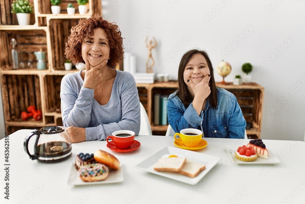 Family of mother and down syndrome daughter sitting at home eating breakfast looking confident at the camera smiling with crossed arms and hand raised on chin. thinking positive. Family of mother and down syndrome daughter sitting at home eating breakfast looking confident at the camera smiling with crossed arms and hand raised on chin. thinking positive.