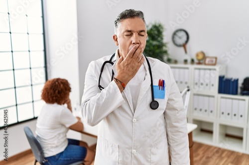 Middle age doctor man at the clinic with a patient bored yawning tired covering mouth with hand. restless and sleepiness.