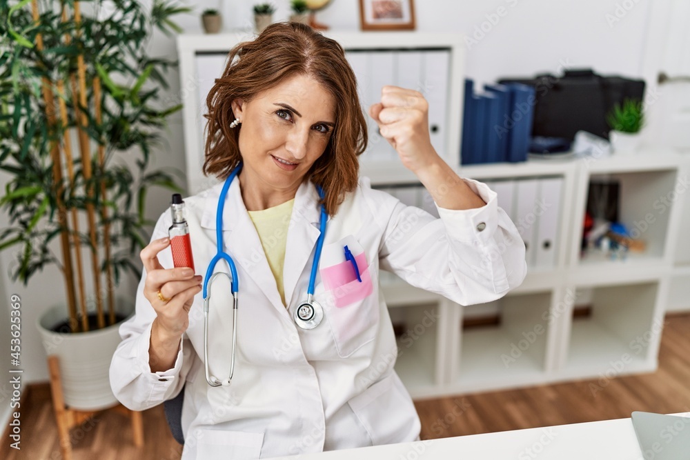 Middle age doctor woman holding electronic cigarette at medical clinic annoyed and frustrated shouting with anger, yelling crazy with anger and hand raised Middle age doctor woman holding electronic cigarette at medical clinic annoyed and frustrated shouting with anger, yelling crazy with anger and hand raised