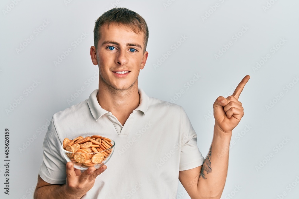 Young caucasian man holding salty biscuits bowl smiling happy pointing with hand and finger to the side Young caucasian man holding salty biscuits bowl smiling happy pointing with hand and finger to the side