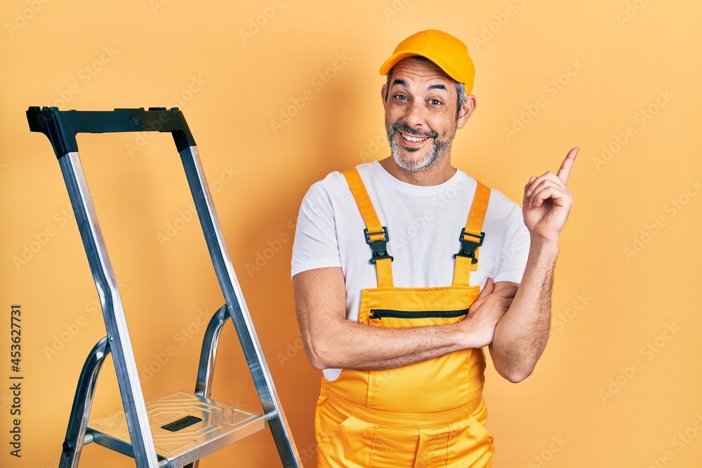 Handsome middle age man with grey hair holding ladder with a big smile on face, pointing with hand and finger to the side looking at the camera.