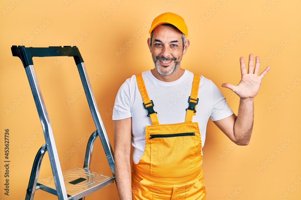 Handsome middle age man with grey hair holding ladder showing and pointing up with fingers number five while smiling confident and happy.