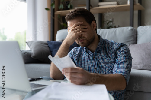 Frustrated desperate millennial man checking bills for payments, holding receipts, getting upset about overspending, too high mortgage, insurance fees. Homeowner analyzing costs, expenses, budget