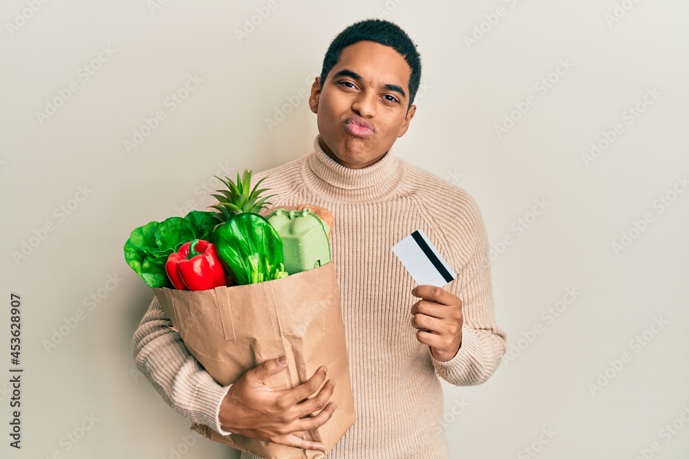 Young handsome hispanic man holding groceries and credit card looking at the camera blowing a kiss being lovely and sexy. love expression. Young handsome hispanic man holding groceries and credit card looking at the camera blowing a kiss being lovely and sexy. love expression.