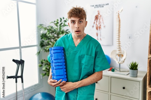 Young caucasian man holding foam roller working at pain recovery clinic scared and amazed with open mouth for surprise  disbelief face