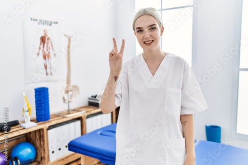 Young caucasian woman working at pain recovery clinic showing and pointing up with fingers number two while smiling confident and happy.