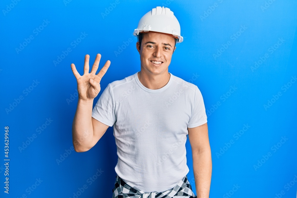 Handsome young man wearing builder uniform and hardhat showing and pointing up with fingers number four while smiling confident and happy.
