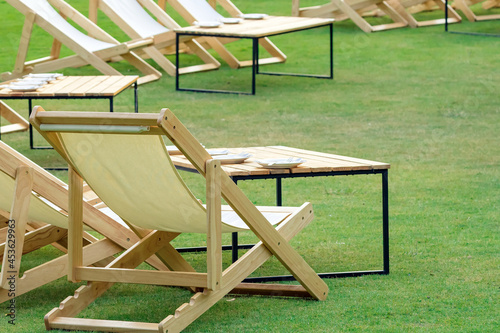 Many empty white deck chairs with tables in lawn is surrounded by shady green grass. Comfortable on outdoor patio chairs in garden.  Lawn chairs in the park. Sunbeds in the garden. Selective focus.