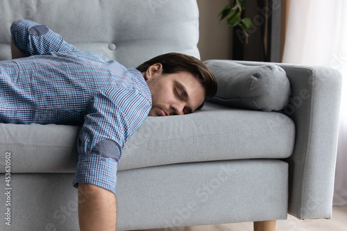 Tired millennial guy in casual sleeping at daytime due to overwork, fatigue, stress, sleepless night. Lazy sleepy bored guy lying on couch with closed eyes, relaxing at home. Energy recreation concept photo
