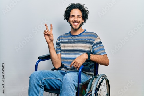 Handsome hispanic man sitting on wheelchair smiling looking to the camera showing fingers doing victory sign. number two.