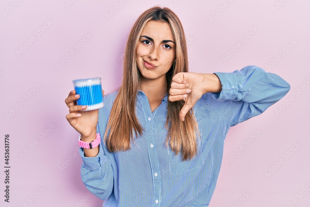 Beautiful hispanic woman holding earwax cotton removers with angry face, negative sign showing dislike with thumbs down, rejection concept Beautiful hispanic woman holding earwax cotton removers with angry face, negative sign showing dislike with thumbs down, rejection concept