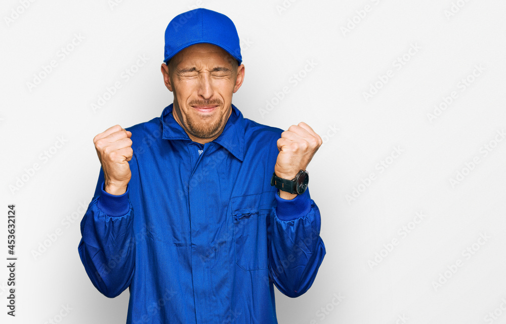Bald man with beard wearing builder jumpsuit uniform excited for success with arms raised and eyes closed celebrating victory smiling. winner concept. Bald man with beard wearing builder jumpsuit uniform excited for success with arms raised and eyes closed celebrating victory smiling. winner concept.