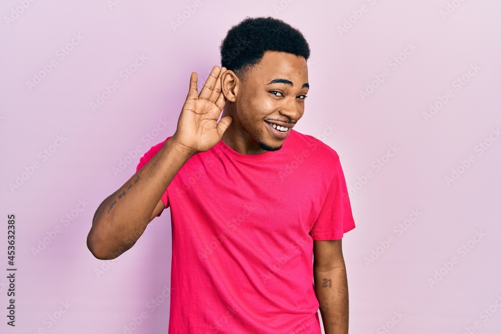 Young african american man wearing casua t shirt smiling with hand over ear listening an hearing to rumor or gossip. deafness concept. Young african american man wearing casua t shirt smiling with hand over ear listening an hearing to rumor or gossip. deafness concept.