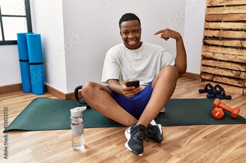 Young african man sitting on training mat at the gym using smartphone looking confident with smile on face  pointing oneself with fingers proud and happy.