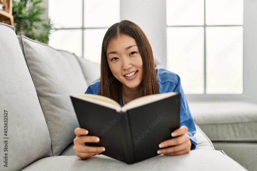 Young chinese girl reading book lying on the sofa at home. Young chinese girl reading book lying on the sofa at home.