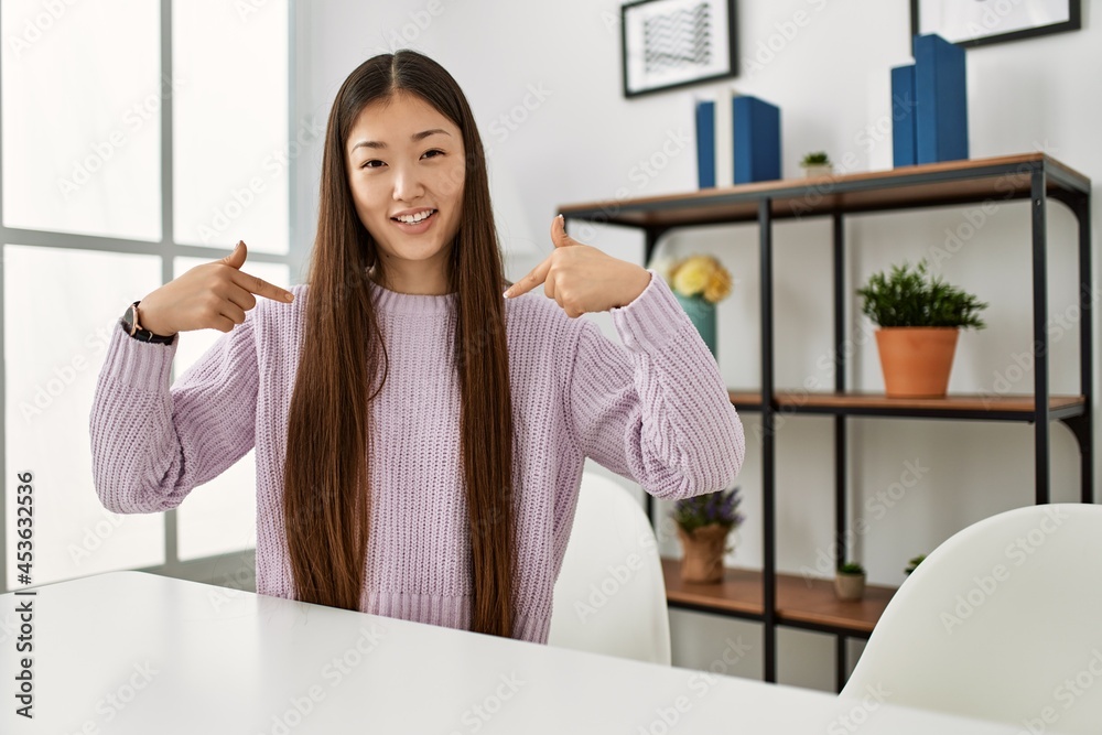 Young chinese girl wearing casual clothes sitting on the table at home looking confident with smile on face, pointing oneself with fingers proud and happy. Young chinese girl wearing casual clothes sitting on the table at home looking confident with smile on face, pointing oneself with fingers proud and happy.