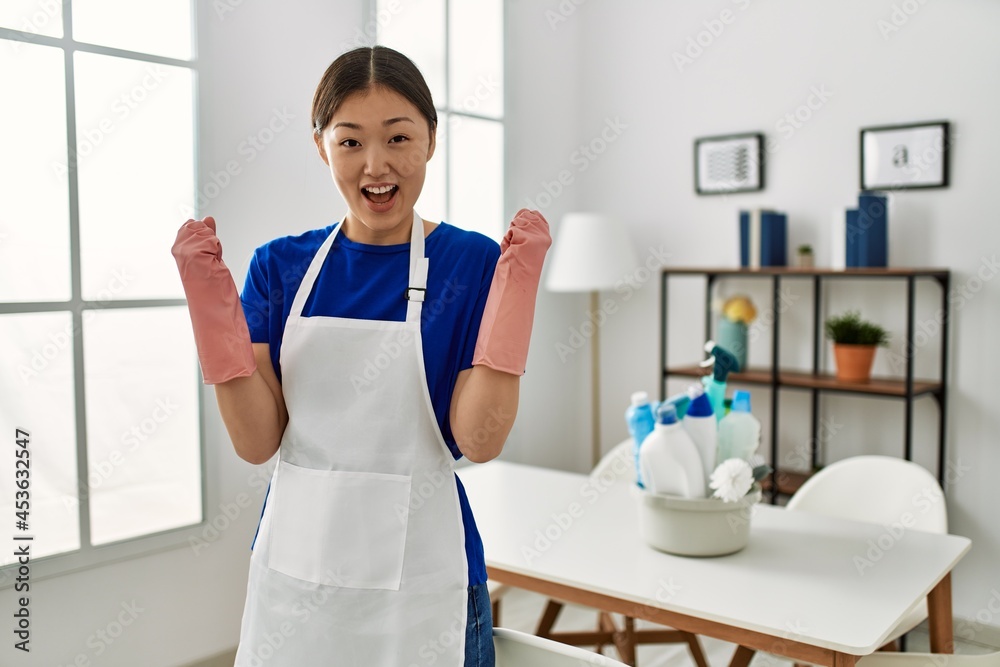 Obraz premium Young chinese girl wearing cleaner uniform standing at home screaming proud, celebrating victory and success very excited with raised arms