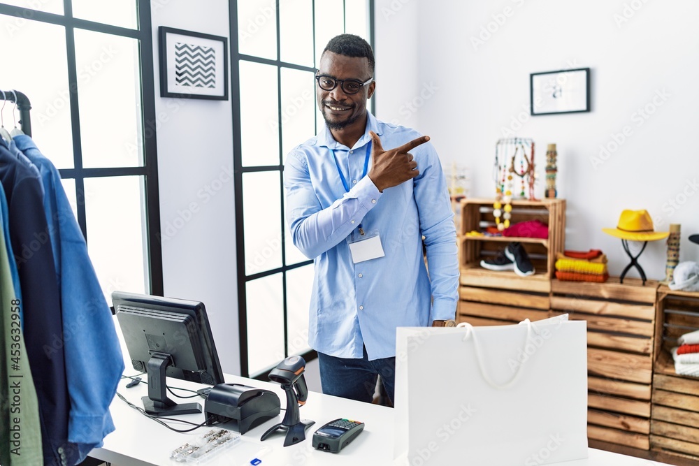 Young african man working as manager at retail boutique cheerful with a smile on face pointing with hand and finger up to the side with happy and natural expression