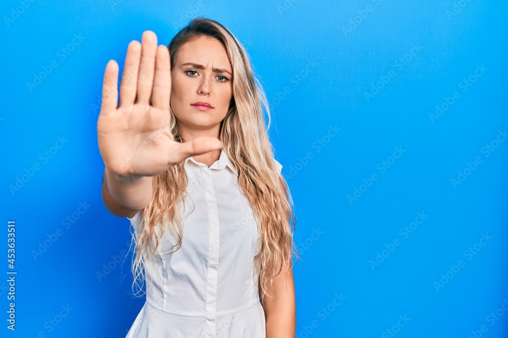 Beautiful young blonde woman wearing casual white shirt doing stop sing with palm of the hand. warning expression with negative and serious gesture on the face.