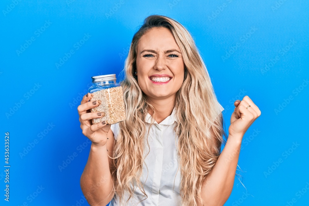 Beautiful young blonde woman holding jar with rice screaming proud, celebrating victory and success very excited with raised arm Beautiful young blonde woman holding jar with rice screaming proud, celebrating victory and success very excited with raised arm
