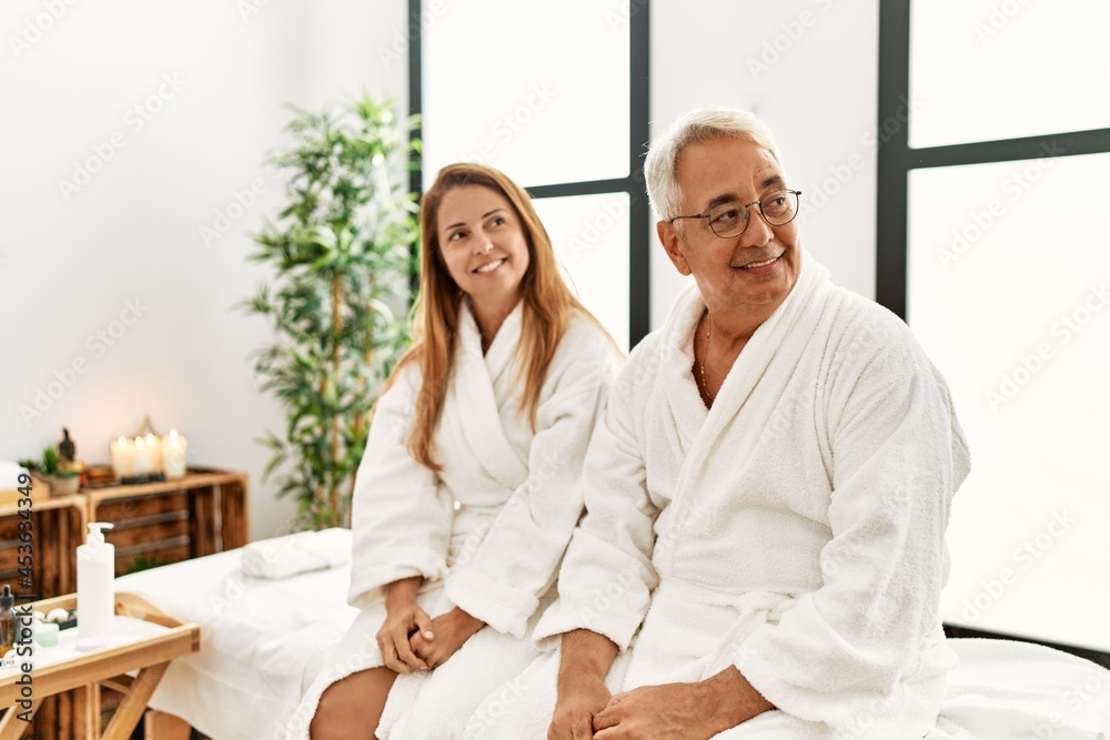 Middle age hispanic couple wearing bathrobe at wellness spa looking away to side with smile on face, natural expression. laughing confident.