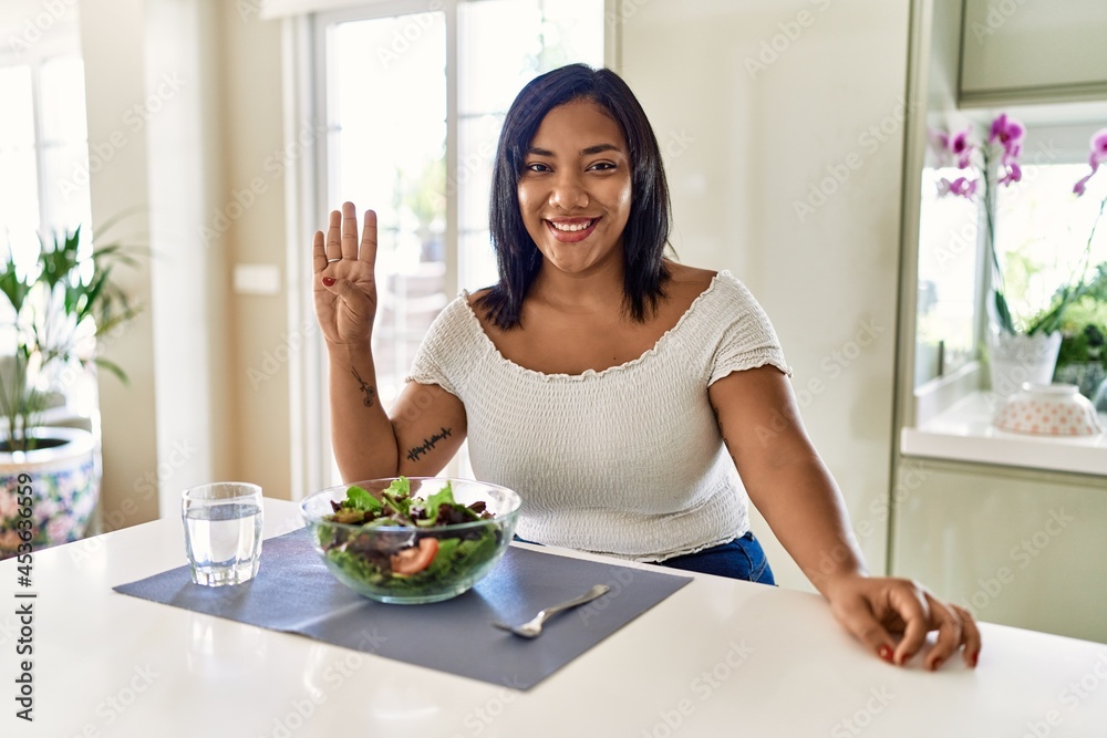 Young hispanic woman eating healthy salad at home showing and pointing up with fingers number four while smiling confident and happy.