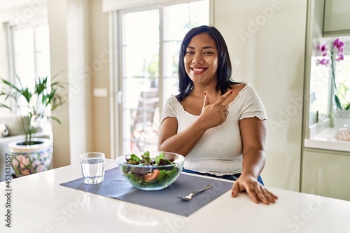 Young hispanic woman eating healthy salad at home cheerful with a smile of face pointing with hand and finger up to the side with happy and natural expression on face