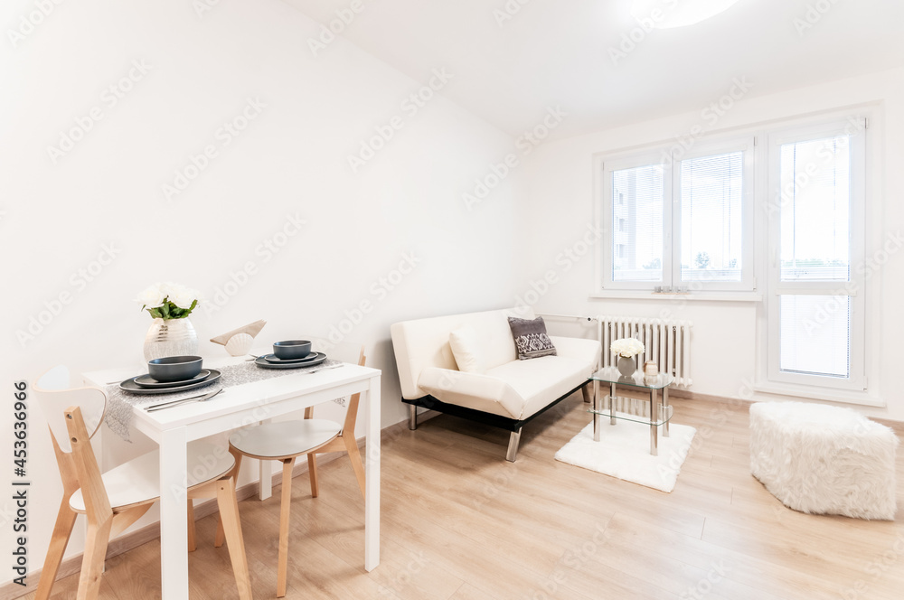 Spacious living room with sofa decorated in modern minimalist style. Wooden materials and white objects predominate in room. At back is a large balcony window. In the foreground a small dining table. Spacious living room with sofa decorated in modern minimalist style. Wooden materials and white objects predominate in room. At back is a large balcony window. In the foreground a small dining table.